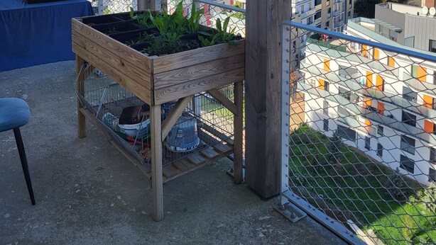 Un balcon agrémenté d’une jardinière en bois contenant des plantes, d’une chaise en métal et offrant une vue sur un paysage urbain avec des bâtiments et une tour.