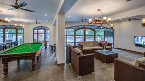 A pool table, comfortable seating, and a television in a spacious room with large windows and ceiling fans. - Villa Calangute Phase 17 (Calangute)