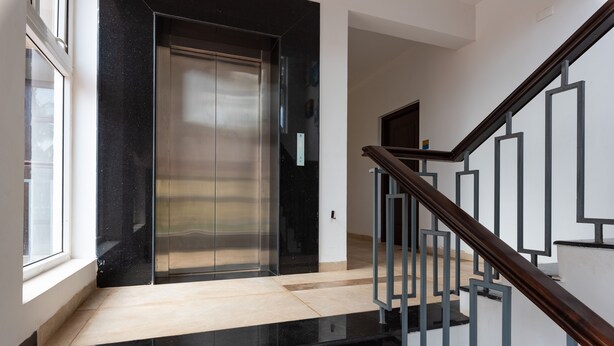 A modern elevator with a metallic finish, a wooden staircase with metal railings, and a light-colored floor. - Villa Calangute Phase 17 (Calangute)