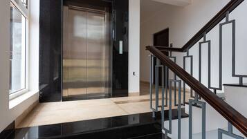 A modern elevator with a metallic finish, a wooden staircase with metal railings, and a light-colored floor.