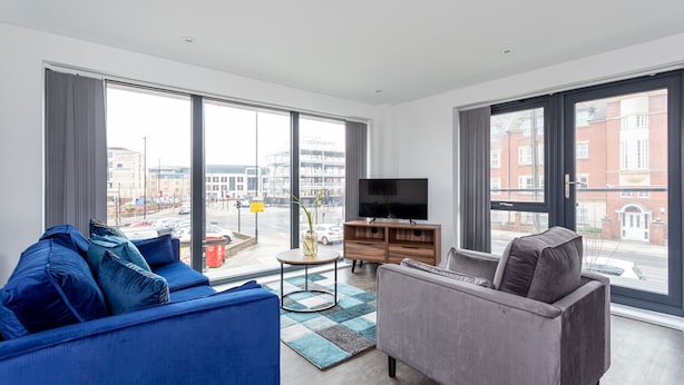 A modern living room with a large window, a flat-screen TV, a blue sofa, a grey sofa, and a wooden cabinet. - Modern & Stylish 2 Bed York Apartment (York)