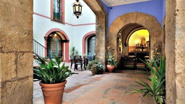 An indoor courtyard with stone flooring, potted plants, and a view of a sunset through an arched doorway.