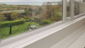 A room with a large window overlooking a rural landscape with fields, trees, and a body of water. - Arfron (Newport)