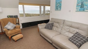 A living room with a beige sofa, a wooden armchair, and a bench. There are two paintings on the wall, a bookshelf, and a view through large windows. - Arfron (Newport)