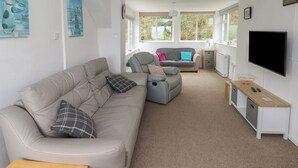 A modern living room with a beige sofa, a grey recliner, and a wooden coffee table. There is a TV on a stand and a small table with a basket. - Arfron (Newport)