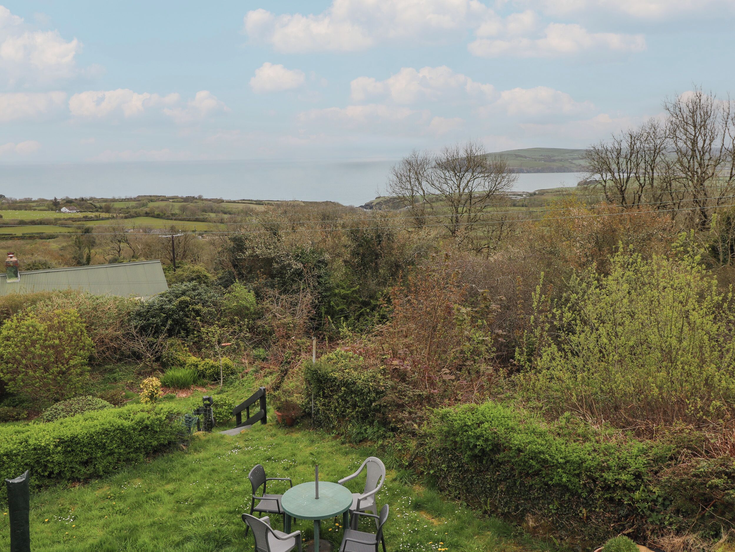 A garden with a table and chairs, surrounded by greenery and a view of the sea.