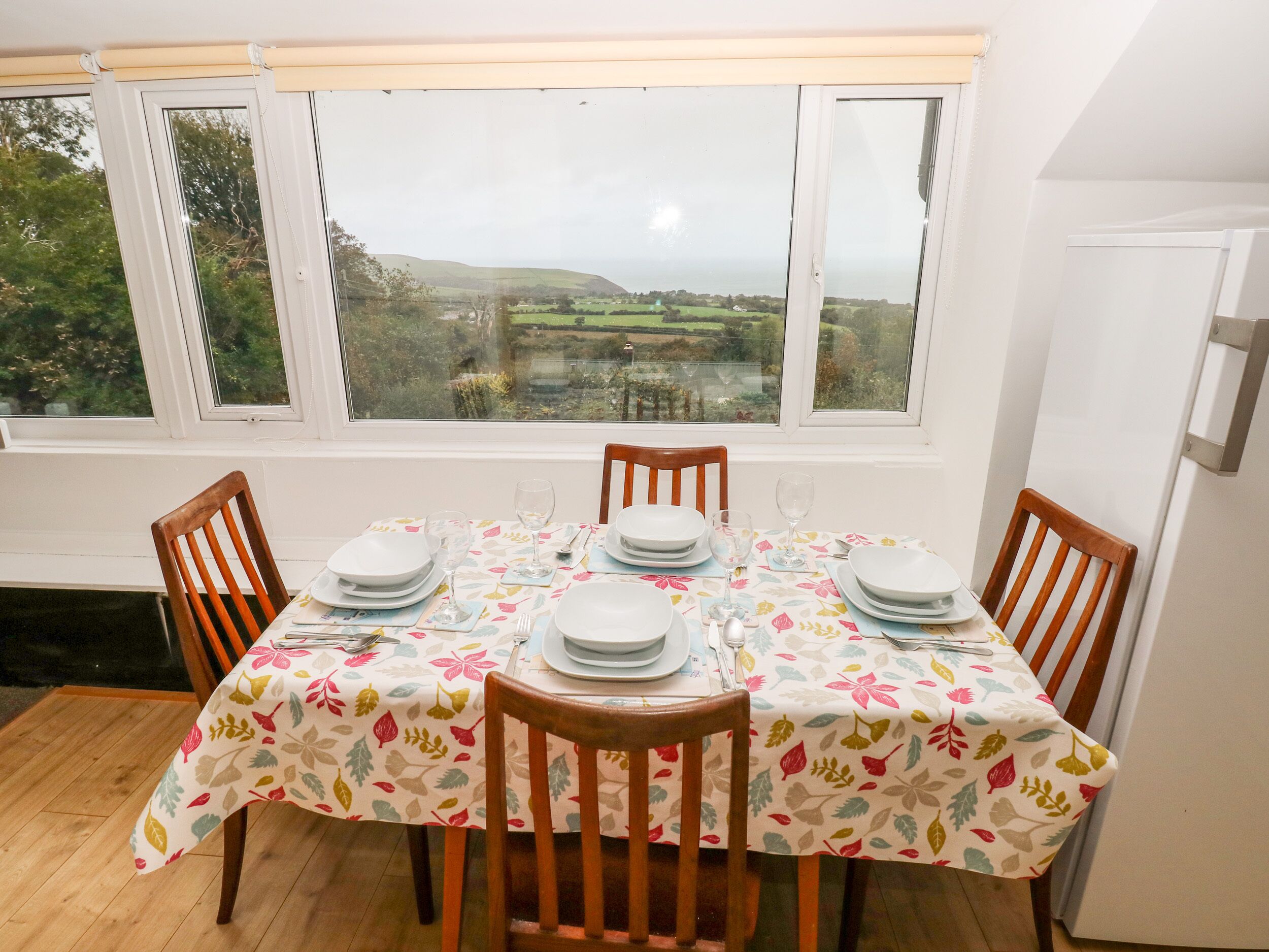 A dining table set for four with a floral tablecloth, positioned by a window with a view of a rural landscape.
