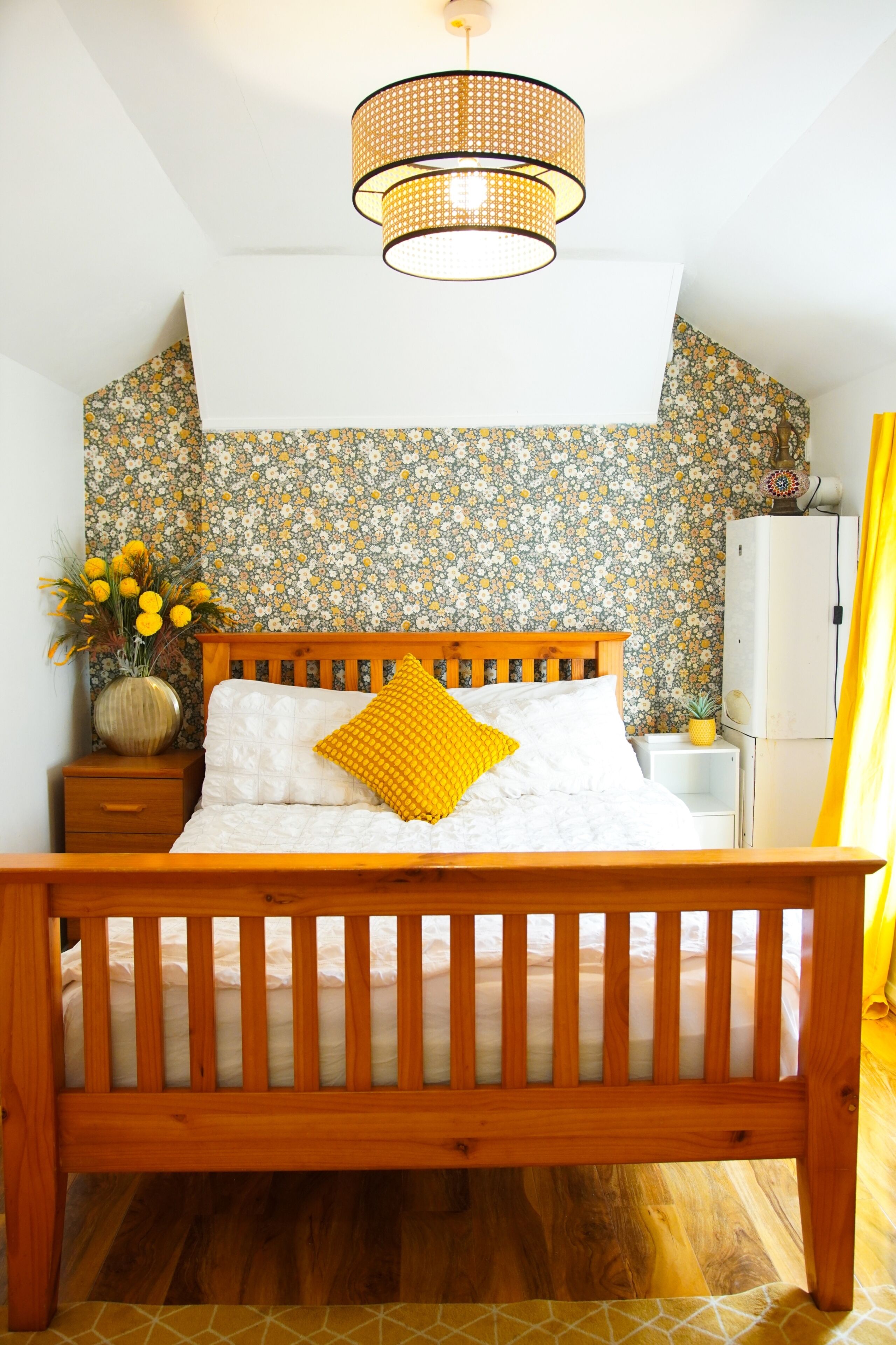 A bedroom with a wooden bed, a nightstand, a vase with flowers, and a floral-patterned wallpaper.