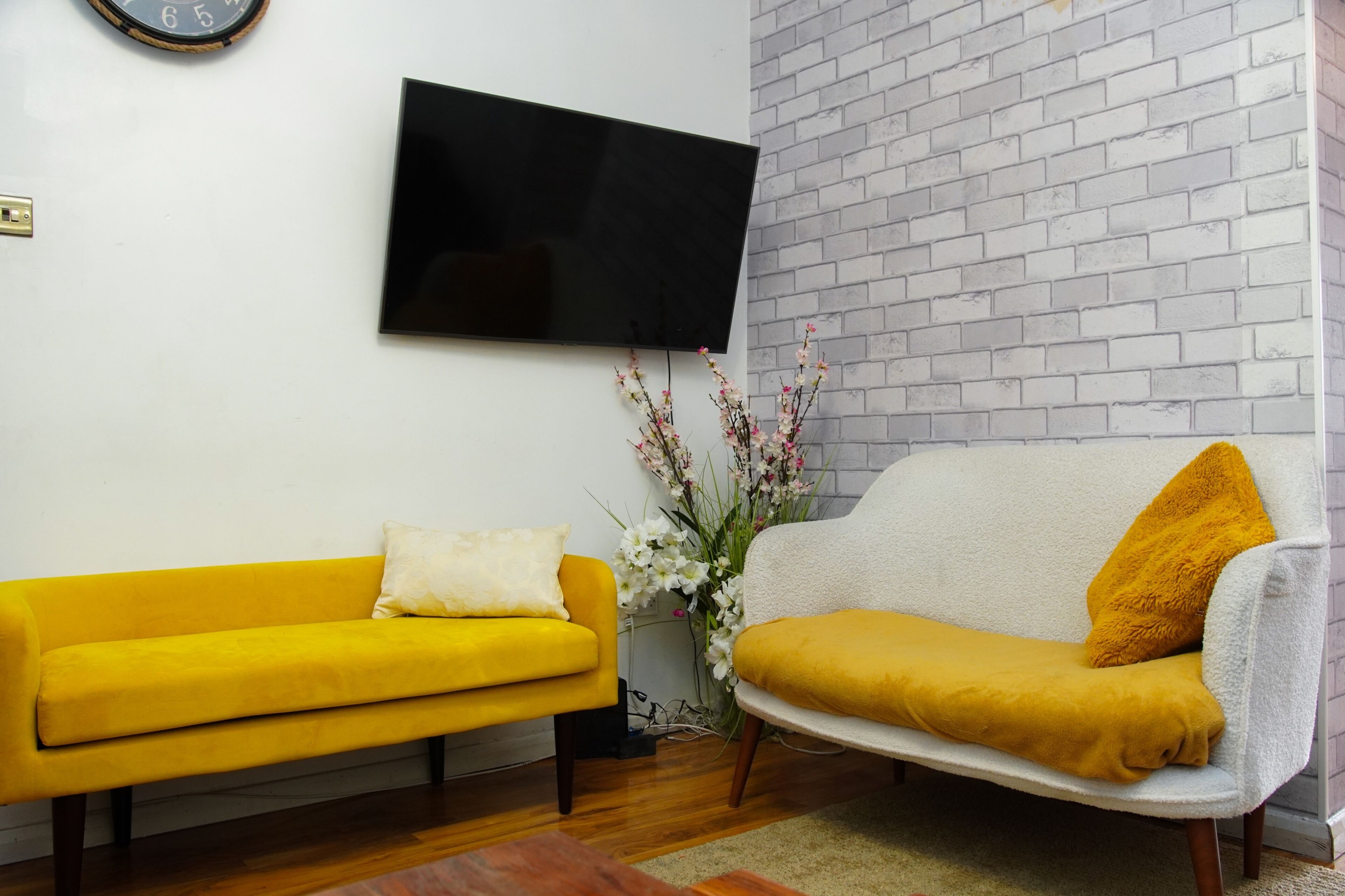 Una sala de estar con un sofá amarillo, un sofá blanco, un televisor de pantalla plana y una pared con diseño de ladrillo.