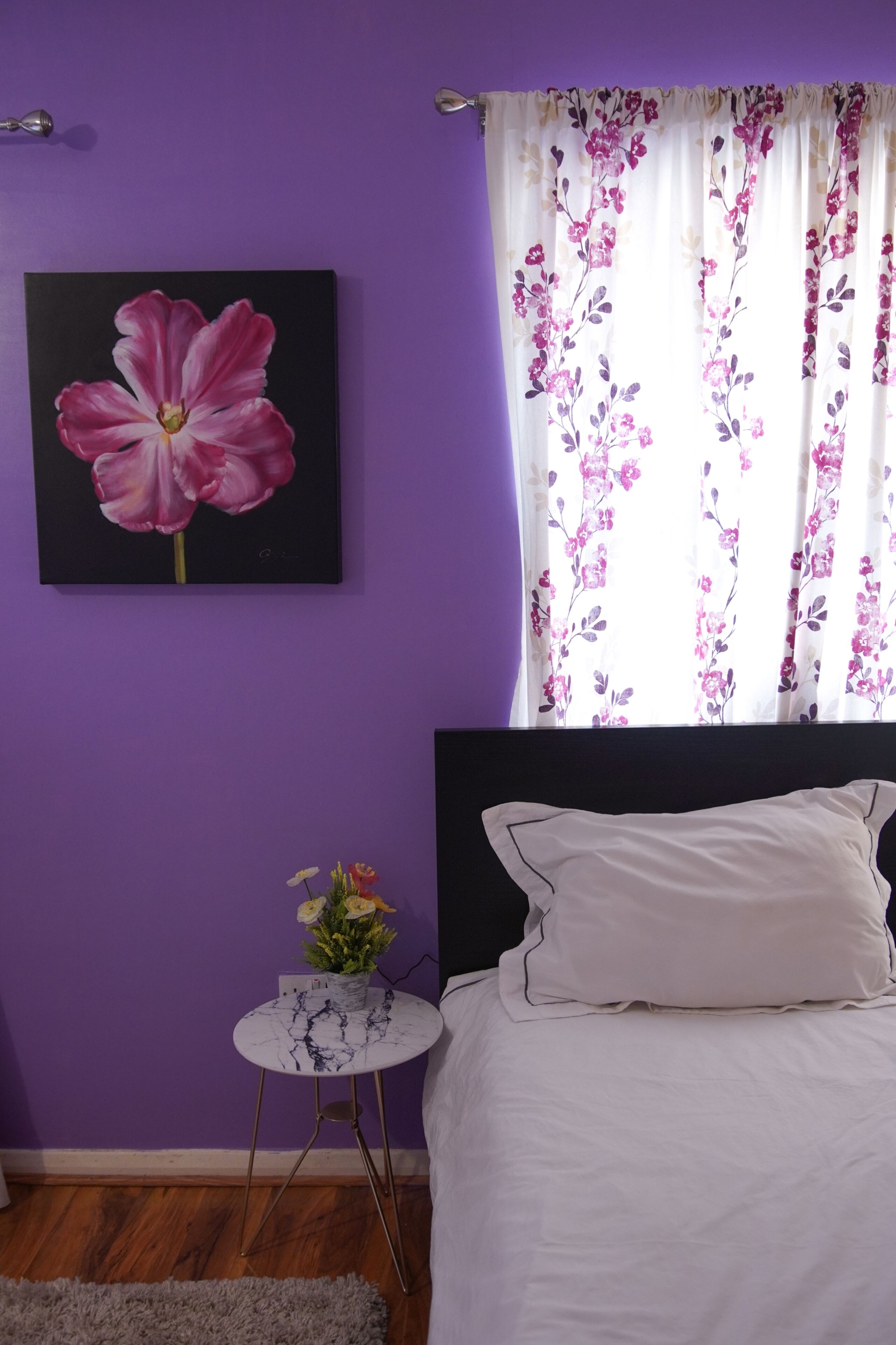 A bedroom with a purple wall, a large floral painting, a bed with white bedding, a small round table with a vase of flowers, and a window with floral curtains.