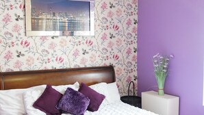 Een slaapkamer met een houten bed, witte beddengoed, paarse kussens, bloemenbehang, een stadsgezicht foto en een vaas met bloemen.