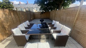 Een overdekt terras buiten met een rechthoekige tafel en acht stoelen, omringd door een houten omheining.