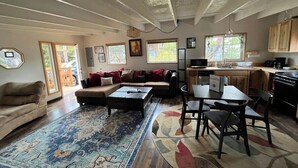 A spacious living room with a sofa, coffee table, and dining area. There are multiple windows providing natural light and a view of the outdoors. - The Chillton Cozy cabin steps to beach (Cloverdale)