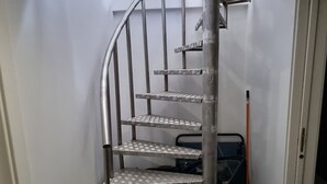 Una escalera de caracol con barandillas metálicas y peldaños con diseño, que conduce a una zona de descanso con una escoba y un objeto pequeño en el suelo.