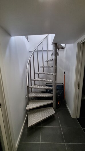 Una escalera de caracol con barandillas metálicas y peldaños con diseño, que conduce a una zona de descanso con una escoba y un objeto pequeño en el suelo.