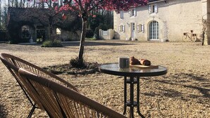 Ein Korbstuhl und ein Tisch mit einem Croissant und einer Tasse in einem Innenhof mit einem Steinhaus und einem Baum mit rosa Blüten.