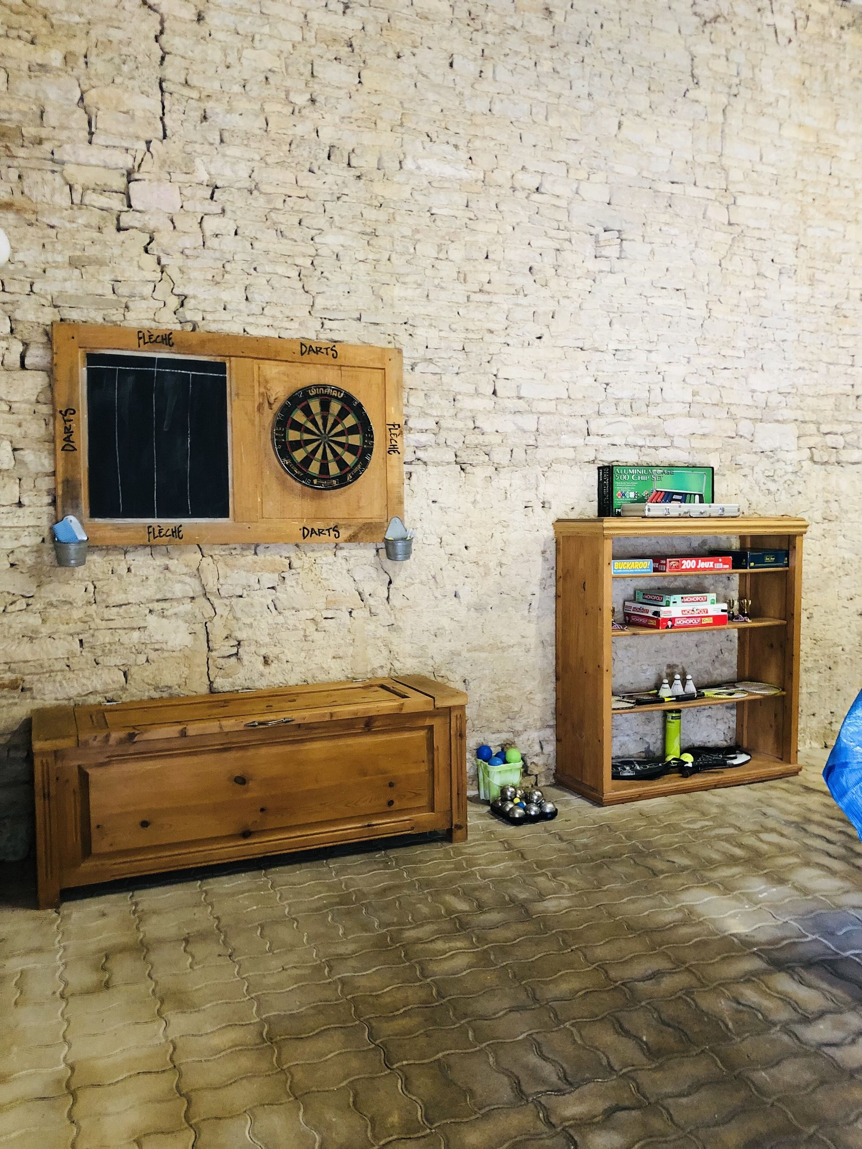 Una habitación con pared de ladrillo, un dardo, un cofre de madera y una estantería con juegos de mesa.