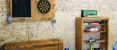 Una habitación con pared de ladrillo, un dardo, un cofre de madera y una estantería con juegos de mesa.