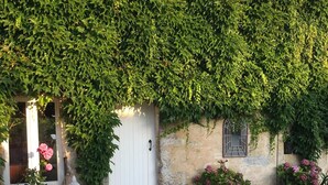 Ein Steinhaus mit einer weißen Tür, umgeben von einer Schotterauffahrt und einem Garten mit rosa Blumen und einer Buddha-Statue.