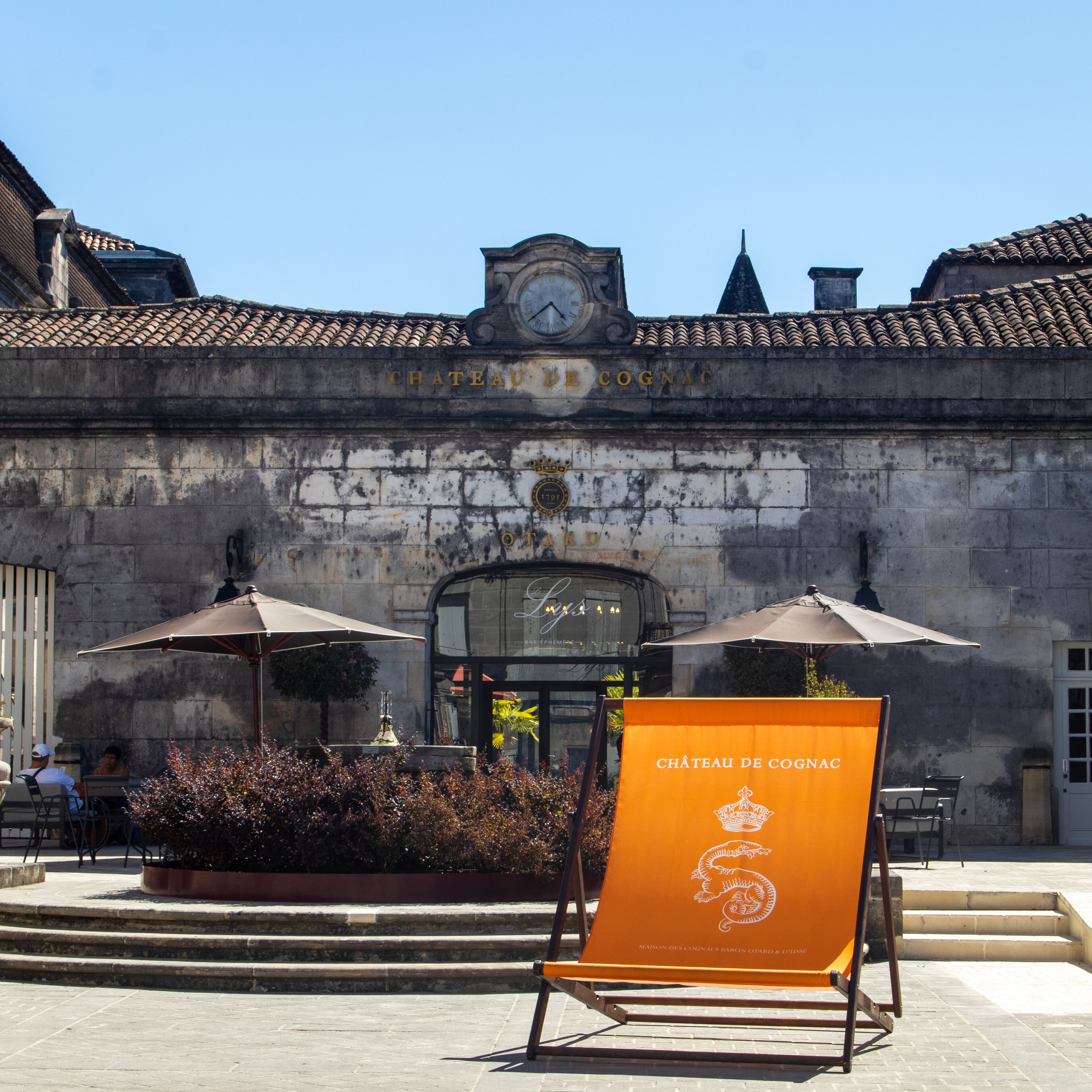 A historic building with a clock tower, outdoor seating with umbrellas, and a sign for Chateau de Cognac.