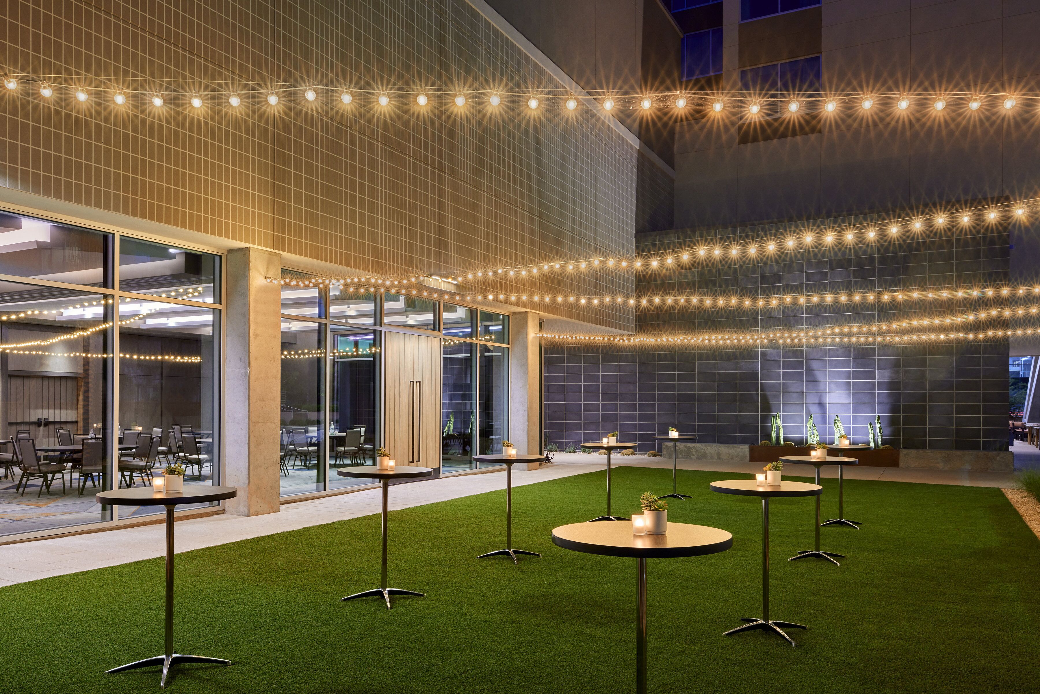 An indoor courtyard with artificial grass, round tables, and string lights.