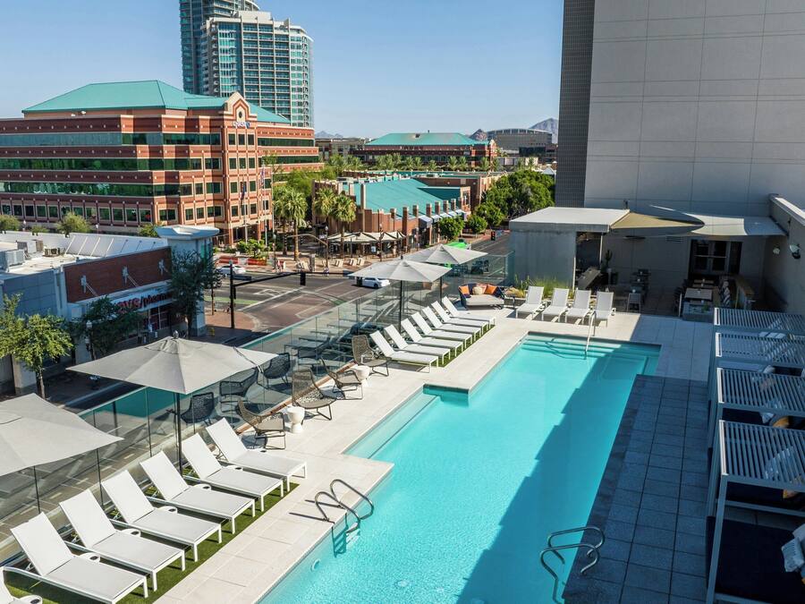 A rooftop pool with lounge chairs and umbrellas, surrounded by buildings and a clear sky.