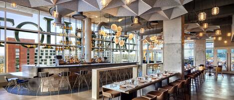 A modern bar area with a long counter, multiple stools, and hanging pendant lights.