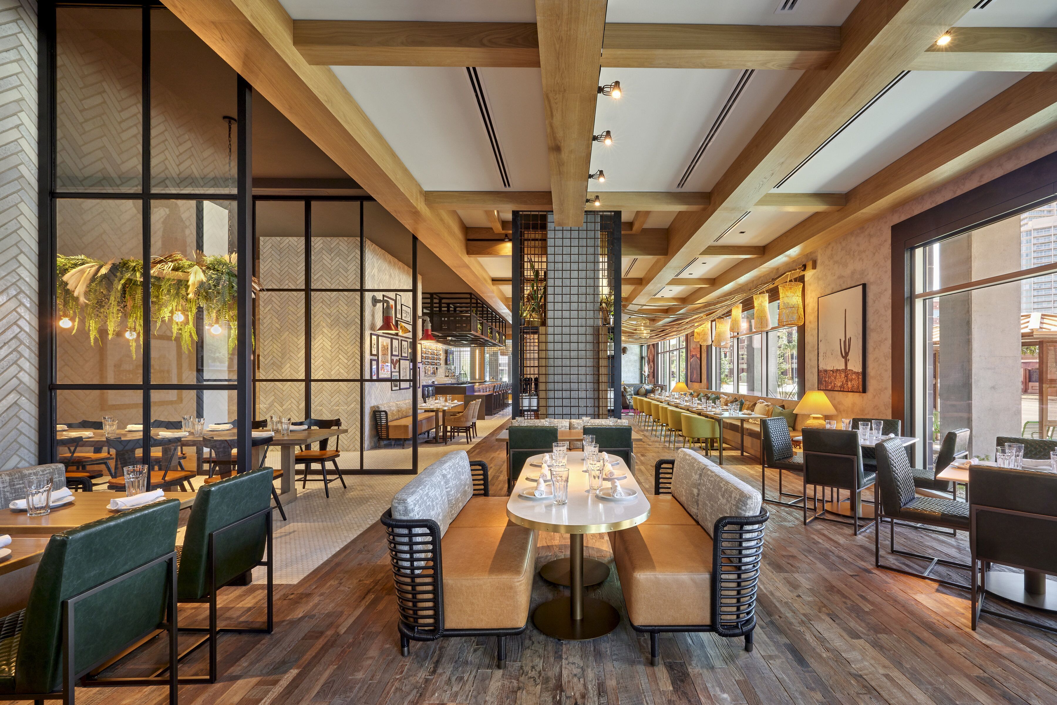 A spacious restaurant with high ceilings, wooden beams, and large windows offering views of the city. Tables are set with white tablecloths and green upholstered chairs.