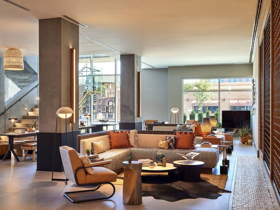 A spacious lobby with a modern sofa, a wooden coffee table, and a seating area with a chair. There is a staircase in the background and a large window providing a view of the outside.