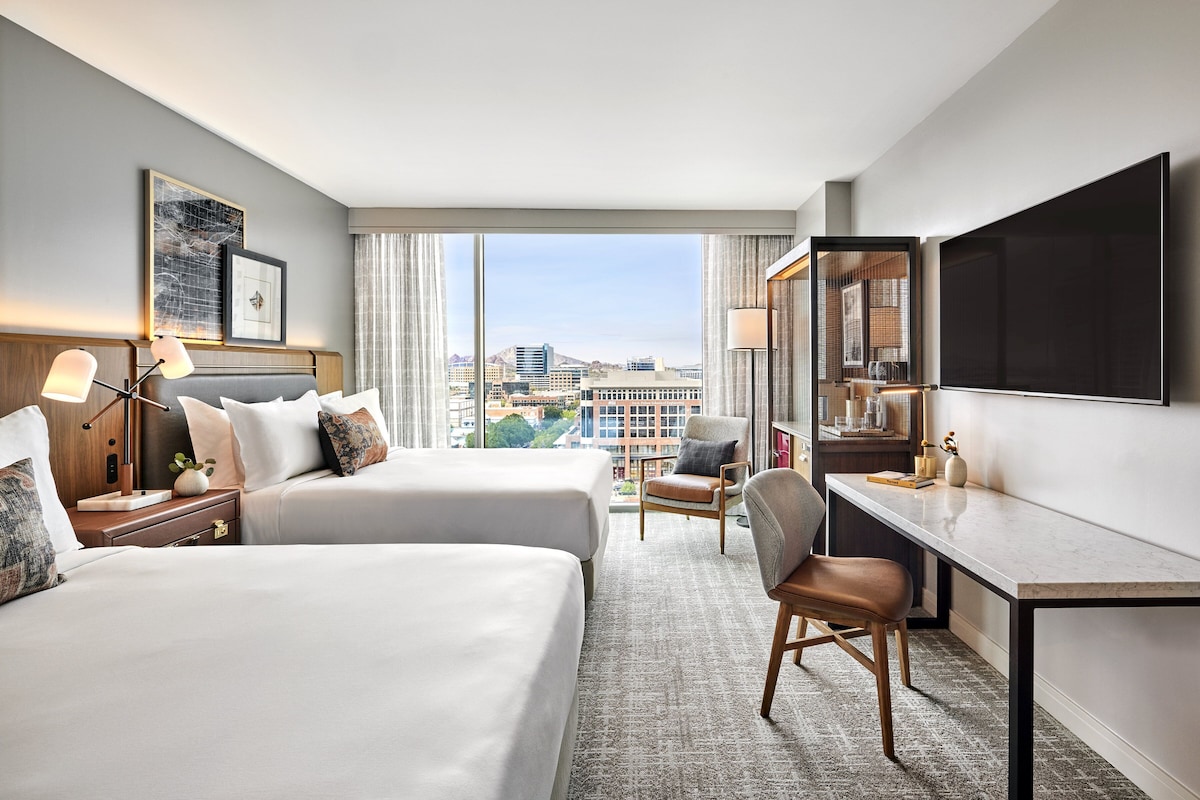 A hotel room with two beds, a desk, a chair, a TV, and a large window offering a city view.