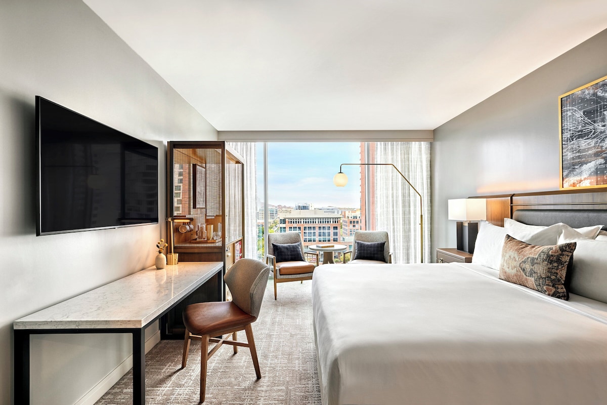 A hotel room with a large bed, a desk, a chair, a TV, and a view of the city.