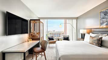 A hotel room with a large bed, a desk, a chair, a TV, and a view of the city.