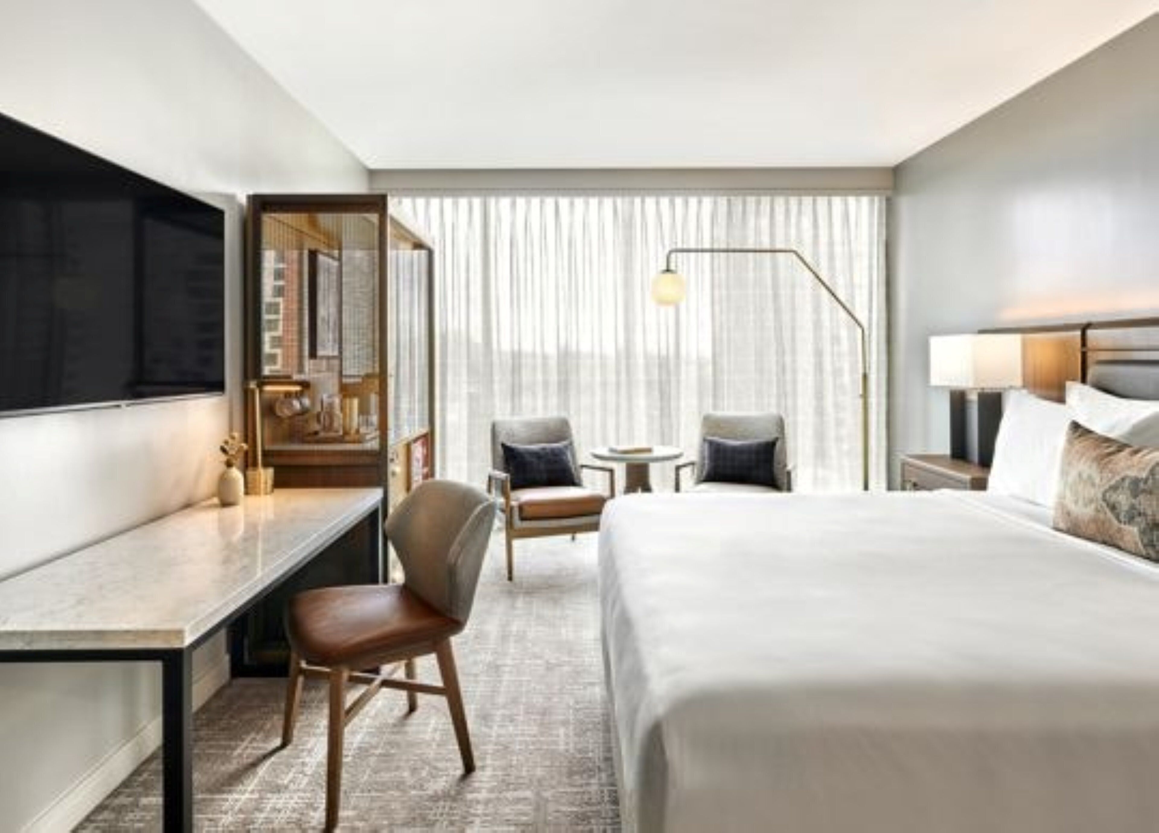 A modern hotel room with a large bed, a desk, a chair, a small table, and a TV.