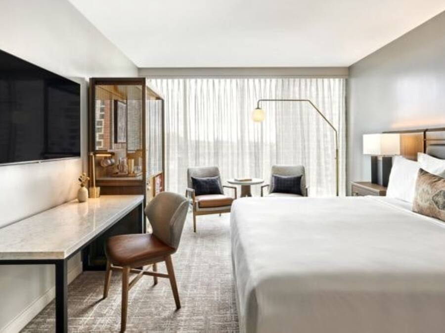 A modern hotel room with a large bed, a desk, a chair, a small table, and a TV.