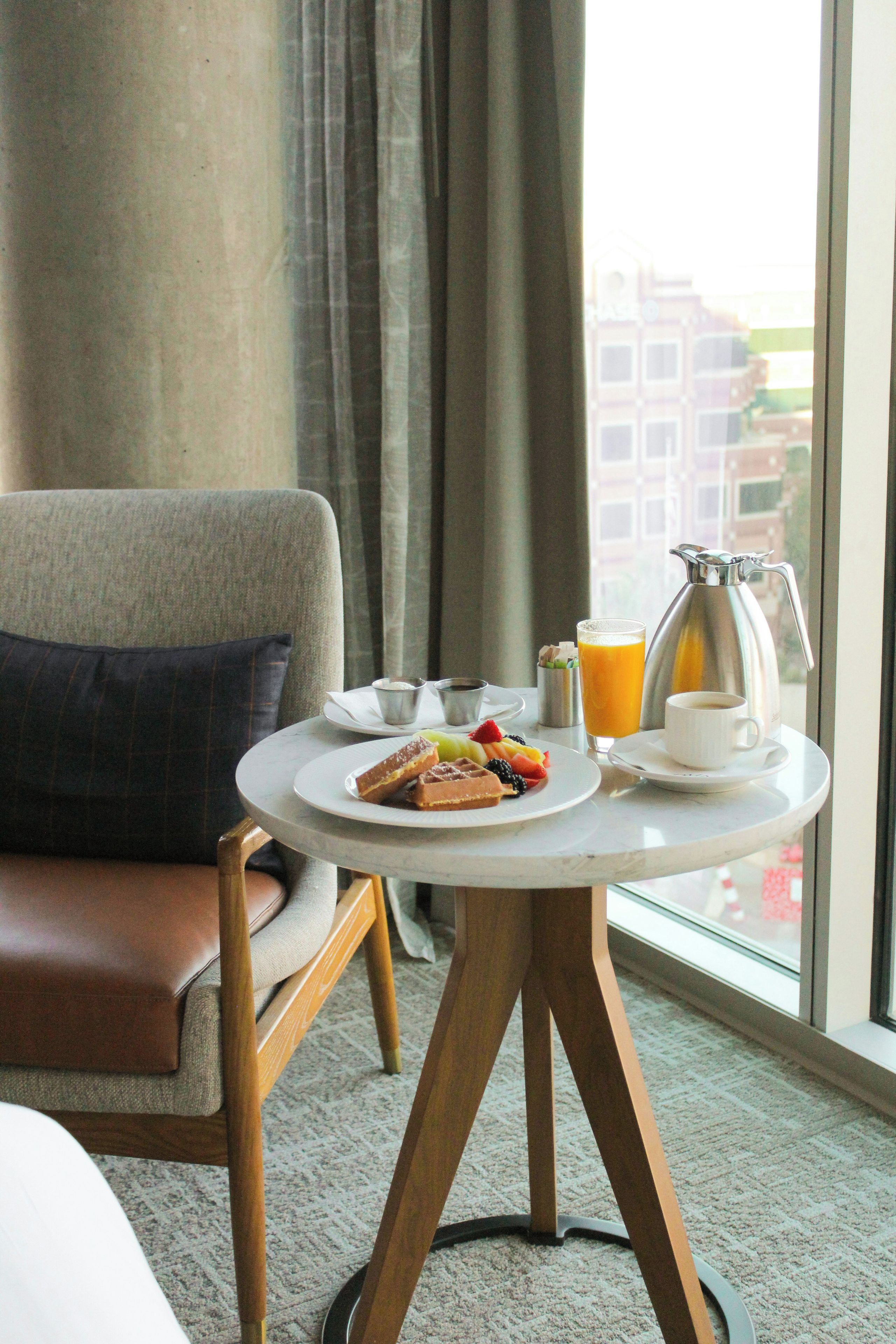 Habitación de hotel con una mesa redonda preparada para el desayuno, que incluye waffles, fruta y una cafetera.