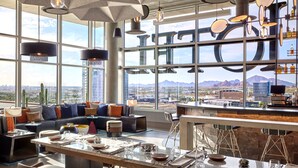 A modern bar area with a panoramic city view, a seating area with a sofa and cushions, and a dining setup with a table set for two. - Omni Tempe Hotel at ASU (Tempe)