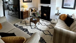 Ein modernes Wohnzimmer mit einer Couch, einem Couchtisch und einem Kamin. Ein Fernseher ist an der Wand montiert und durch einen offenen Durchgang ein Essbereich sichtbar.