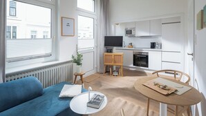 Sala de estar moderna com sofá azul, uma pequena mesa branca e uma mesa de jantar de madeira com cadeiras. Uma televisão está fixada na parede, e há uma área de cozinha com armários e eletrodomésticos brancos.