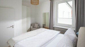 Quarto com cama, uma cadeira, janela com cortinas e radiador.
