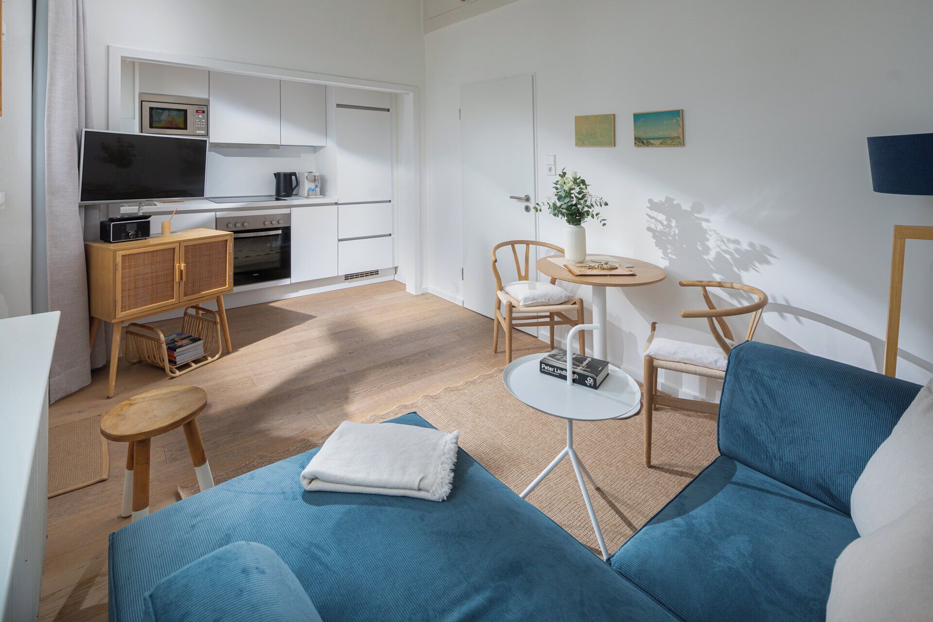 A modern living room with a blue sofa, a small round table, and a kitchen area with white cabinets and a microwave.