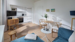 Uma sala de estar moderna com um sofá azul, uma mesa redonda pequena e uma área de cozinha com armários brancos e um micro-ondas.