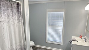 Une salle de bain avec un rideau de douche, des toilettes, une fenêtre avec des stores, un lavabo avec un miroir et deux tapis sur le sol.