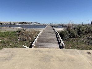 Un chemin en béton mène à une jetée en bois qui s’avance sur une étendue d’eau, avec un pont en arrière-plan.