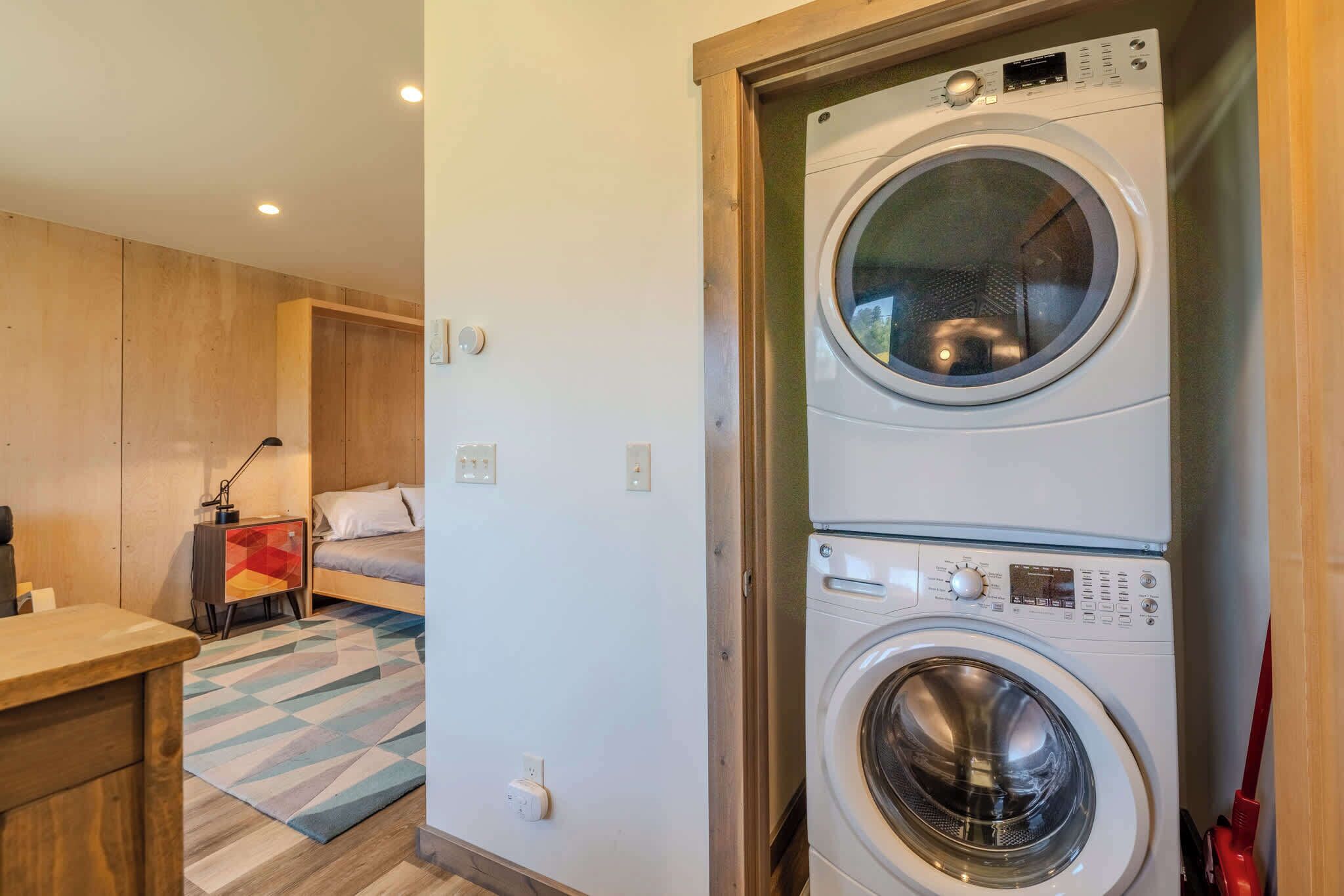 Une buanderie équipée d’un lave-linge et d’un sèche-linge superposés, un lit, un bureau et une table de chevet avec une lampe.
