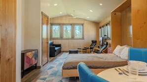 Une chambre d’hôtel moderne dotée d’un grand lit, d’un coin repas, d’un espace salon avec des fauteuils et offrant une vue sur les arbres par les fenêtres.
