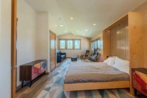 A modern hotel room with a large bed, a seating area with a sofa and chairs, a wooden wardrobe, and a view of the outdoors through the windows. - YCC Studio (West Yellowstone)