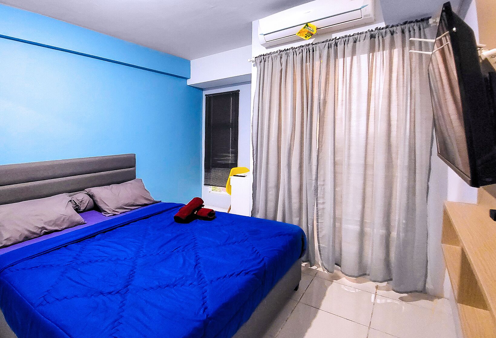 A bedroom with a blue bedspread, grey pillows, and a grey headboard. There is a window with blinds, a curtain, and a TV mounted on the wall.