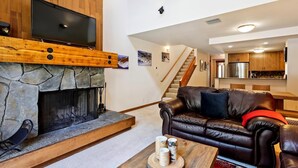 A modern living room with a stone fireplace, a leather sofa, and a flat-screen TV mounted above it. - One Bedroom plus Loft River View Condo (Alpine Meadows)