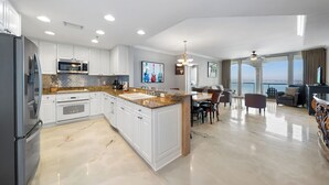 Cozinha moderna com armários brancos, bancadas de granito e eletrodomésticos de aço inoxidável. Possui área de jantar com mesa e cadeiras, e vista para o oceano através de grandes janelas.