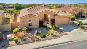 A residential house with a tiled roof, a front entrance with a cross, and a landscaped garden with various plants and a small pool. - Luxury Stay at Estrella Mountain Ranch (Goodyear)